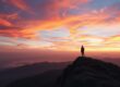 Figura solitária de pé no topo da montanha durante o amanhecer, representando inspiração espiritual e superação pela fé.
